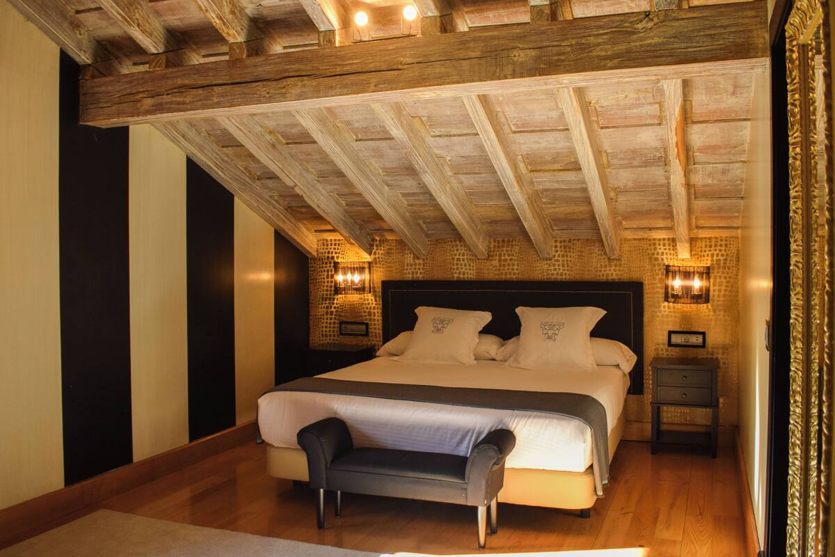 A bedroom with a large bed, wooden ceiling beams, two bedside lamps, and a bench.