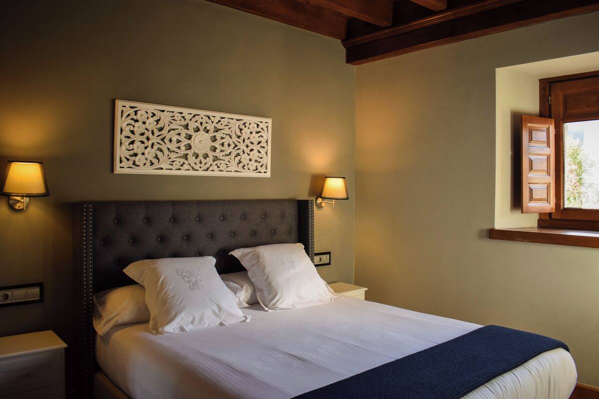 A neatly made bed with white linens and pillows, a decorative wall piece above it, and two wall-mounted lamps on either side.