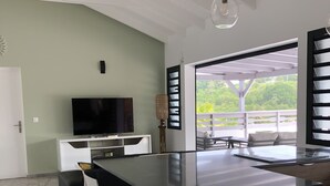 A modern kitchen with a large island, white bar stools, a flat-screen TV, and a view of greenery through large windows. - T4 Sainte Anne - Fonds Thezan (Sainte-Anne)