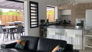 A modern kitchen with white cabinetry, a built-in oven, and a dining area with a black leather sofa and colorful cushions. - T4 Sainte Anne - Fonds Thezan (Sainte-Anne)