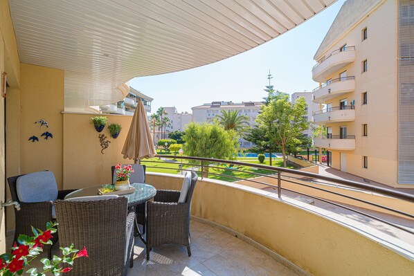 A balcony with wicker furniture, potted plants, and a view of a landscaped garden and buildings.