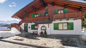 Een houten chalet met balkon, een stenen trap en een besneeuwde berg op de achtergrond.