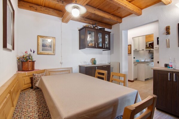 Une pièce avec un plafond en bois, une table à manger recouverte d’une nappe, un coin cuisine avec des placards et un tableau encadré accroché au mur.