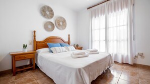 A neatly made bed with white linens and blue pillows, a wooden headboard, a bedside table with a plant, and a large window with sheer curtains.