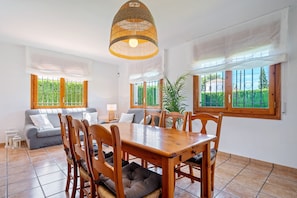 Een eethoek met een houten tafel en stoelen, een hanglamp en grote ramen met uitzicht op groen.