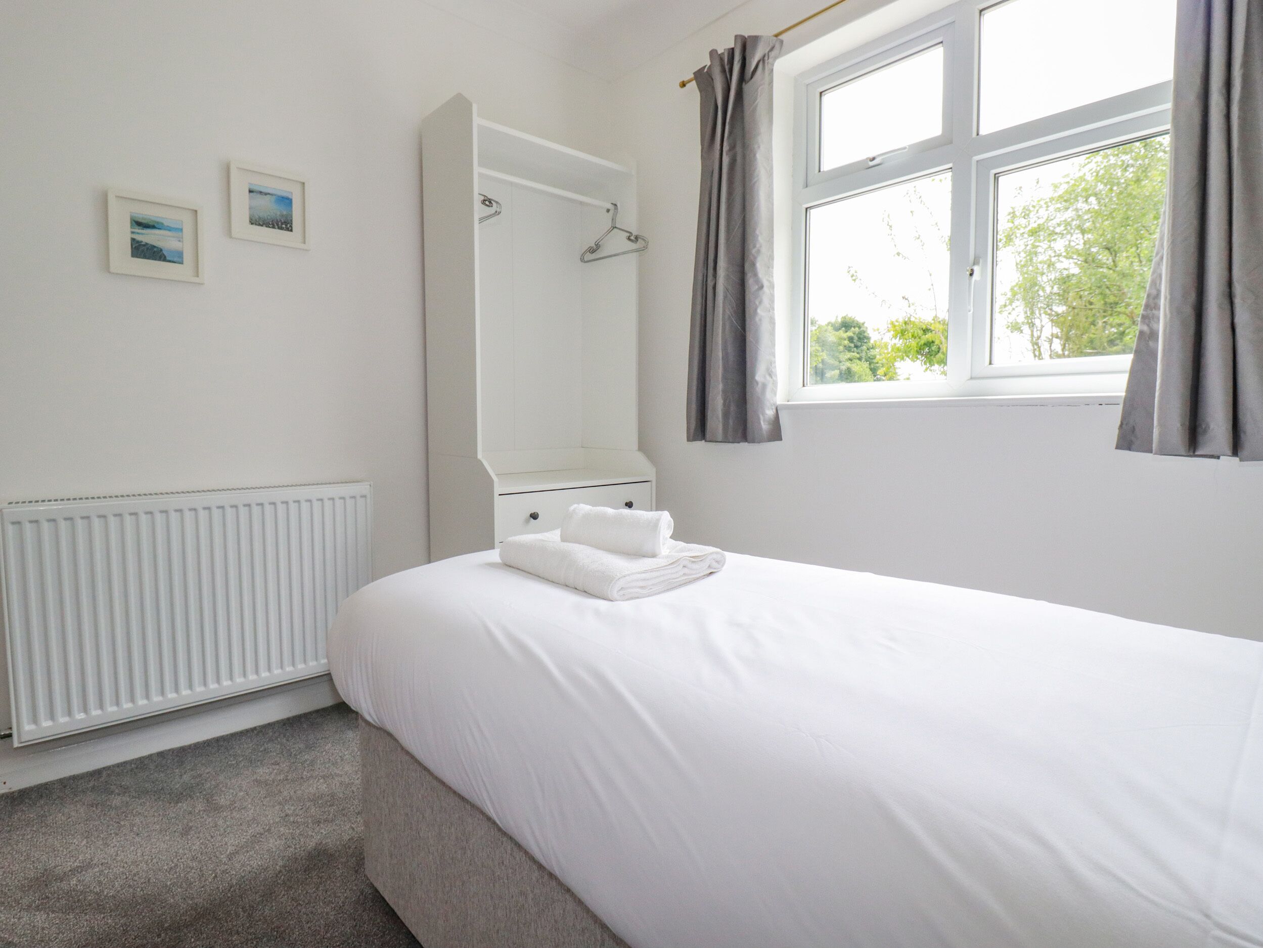 Une chambre à coucher avec un lit, un radiateur, une fenêtre avec des rideaux et deux tableaux encadrés accrochés au mur.