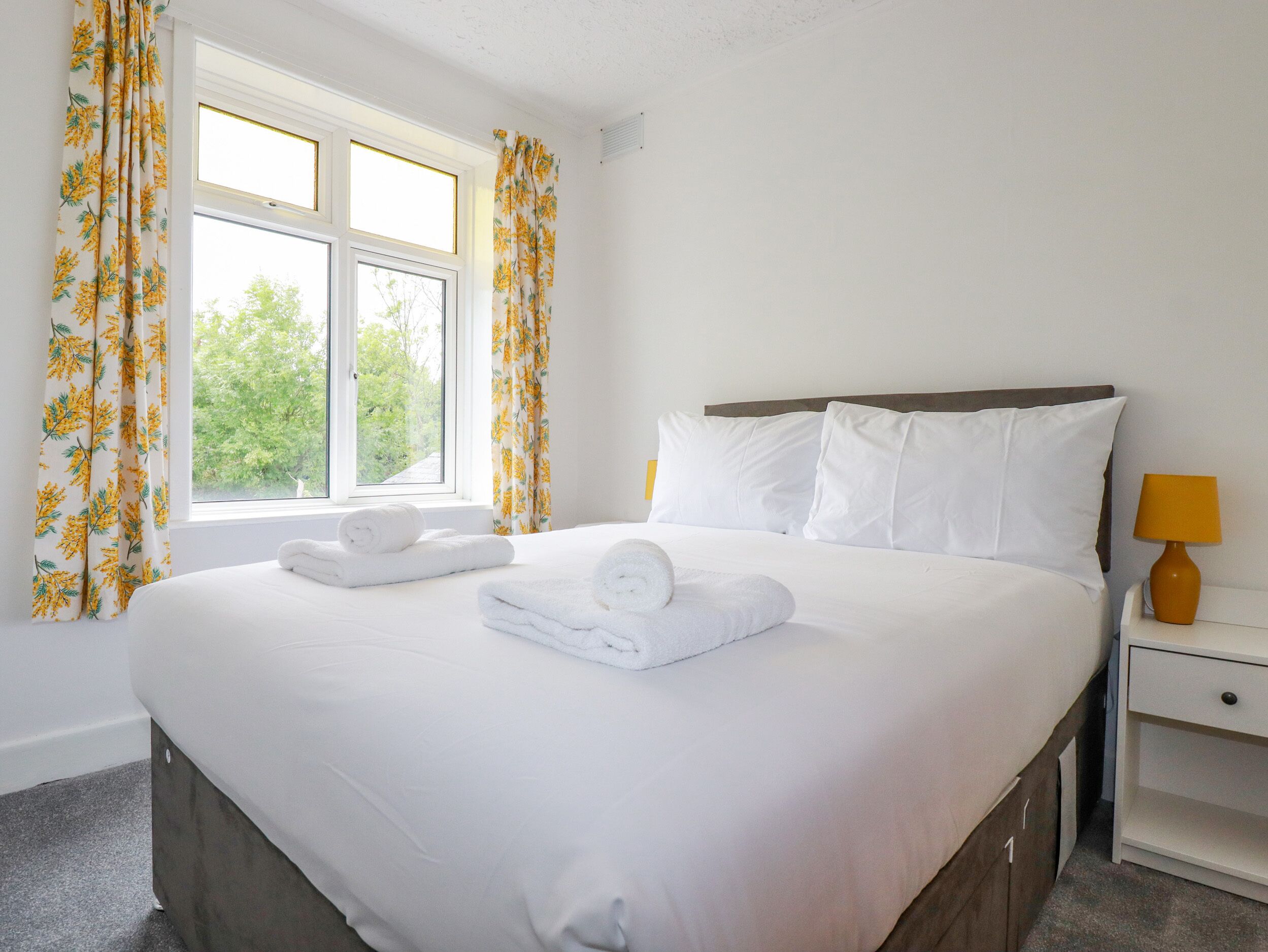 A neatly made bed with white linens and pillows, a window with floral curtains, and a bedside table with a lamp.
