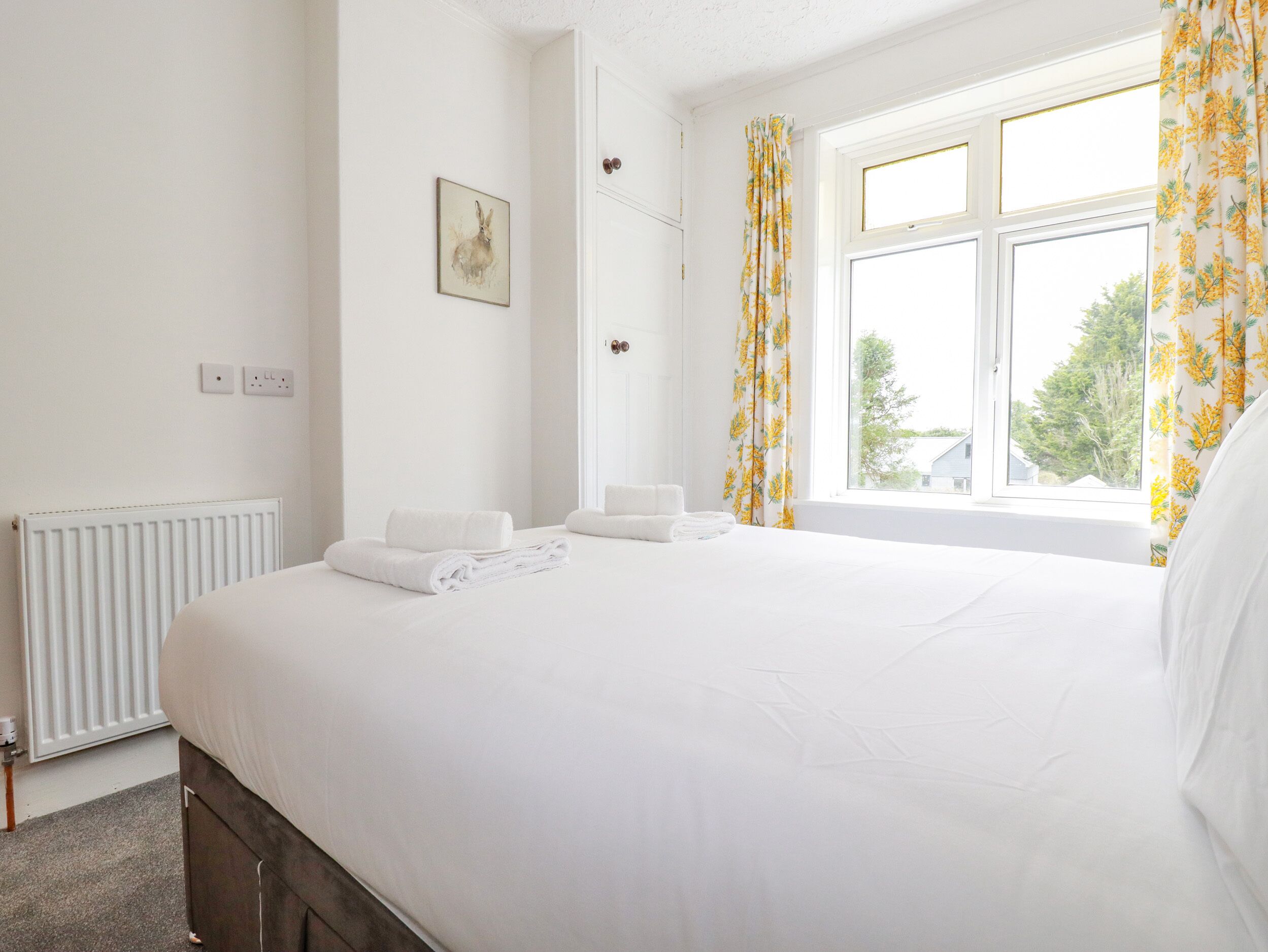 A neatly made bed with white linens, a radiator, a framed picture on the wall, and a window with floral curtains.