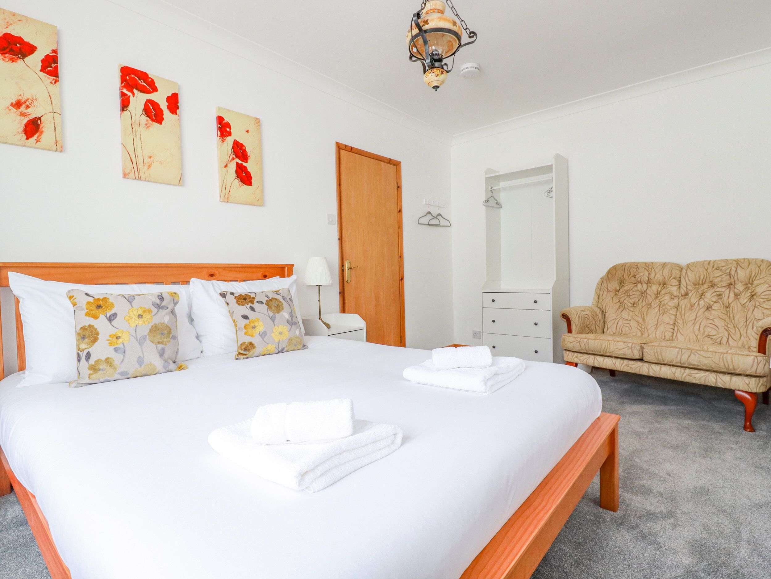 A neatly made bed with white linens and floral pillows, a wooden headboard, a beige sofa, a wooden nightstand with a lamp, and a mirror on the wall.