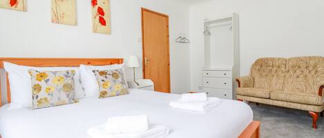 A neatly made bed with white linens and floral pillows, a wooden headboard, a beige sofa, a wooden nightstand with a lamp, and a mirror on the wall.