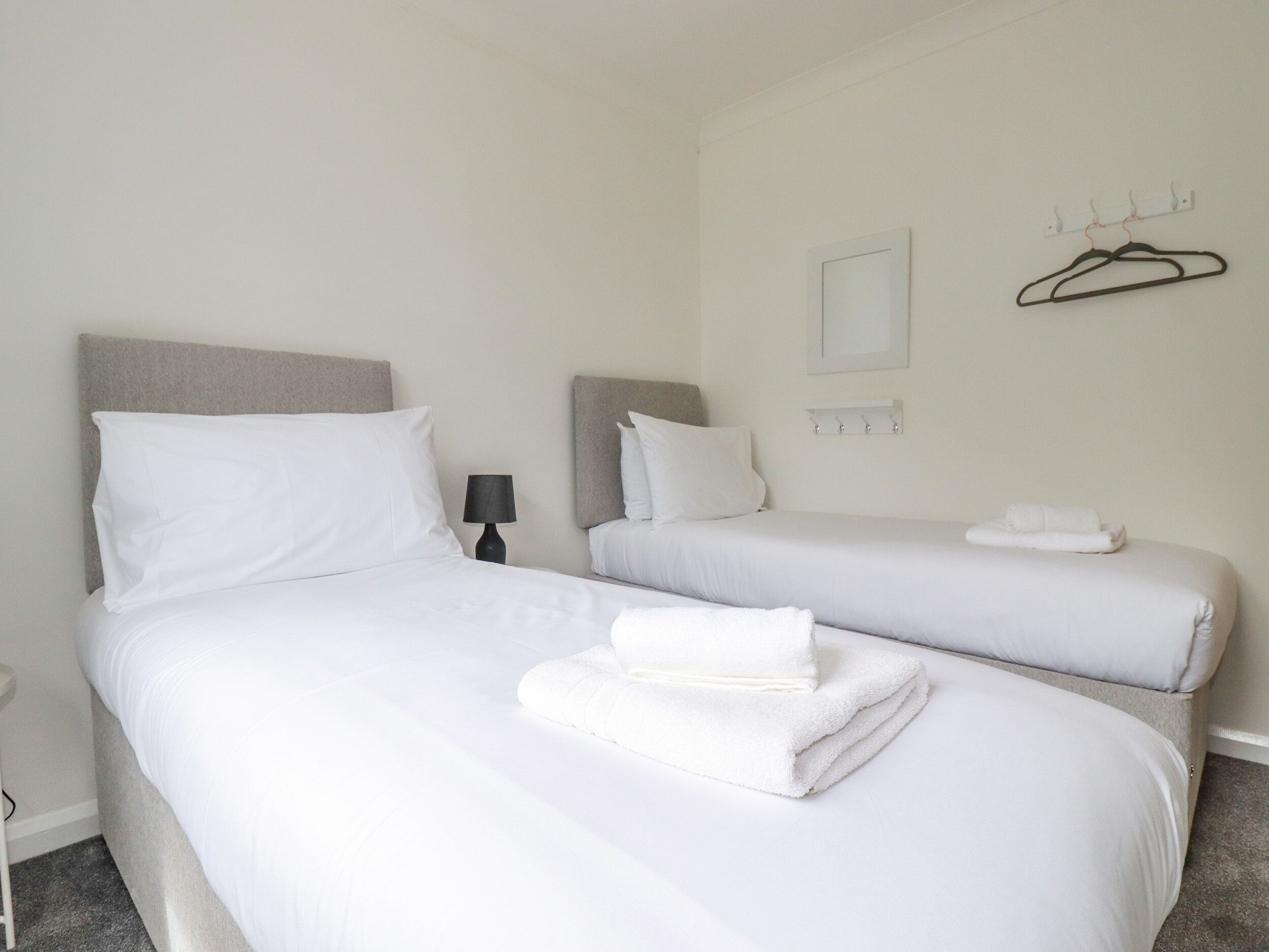 Een hotelkamer met twee bedden, witte lakens, een bedlampje, een kapstok en een lijst op de muur.