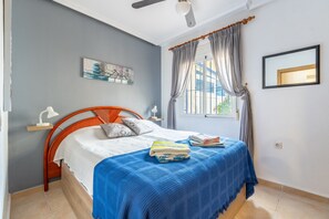 Un dormitorio con cama, ventana con cortinas, ventilador de techo, espejo en la pared y un cuadro.