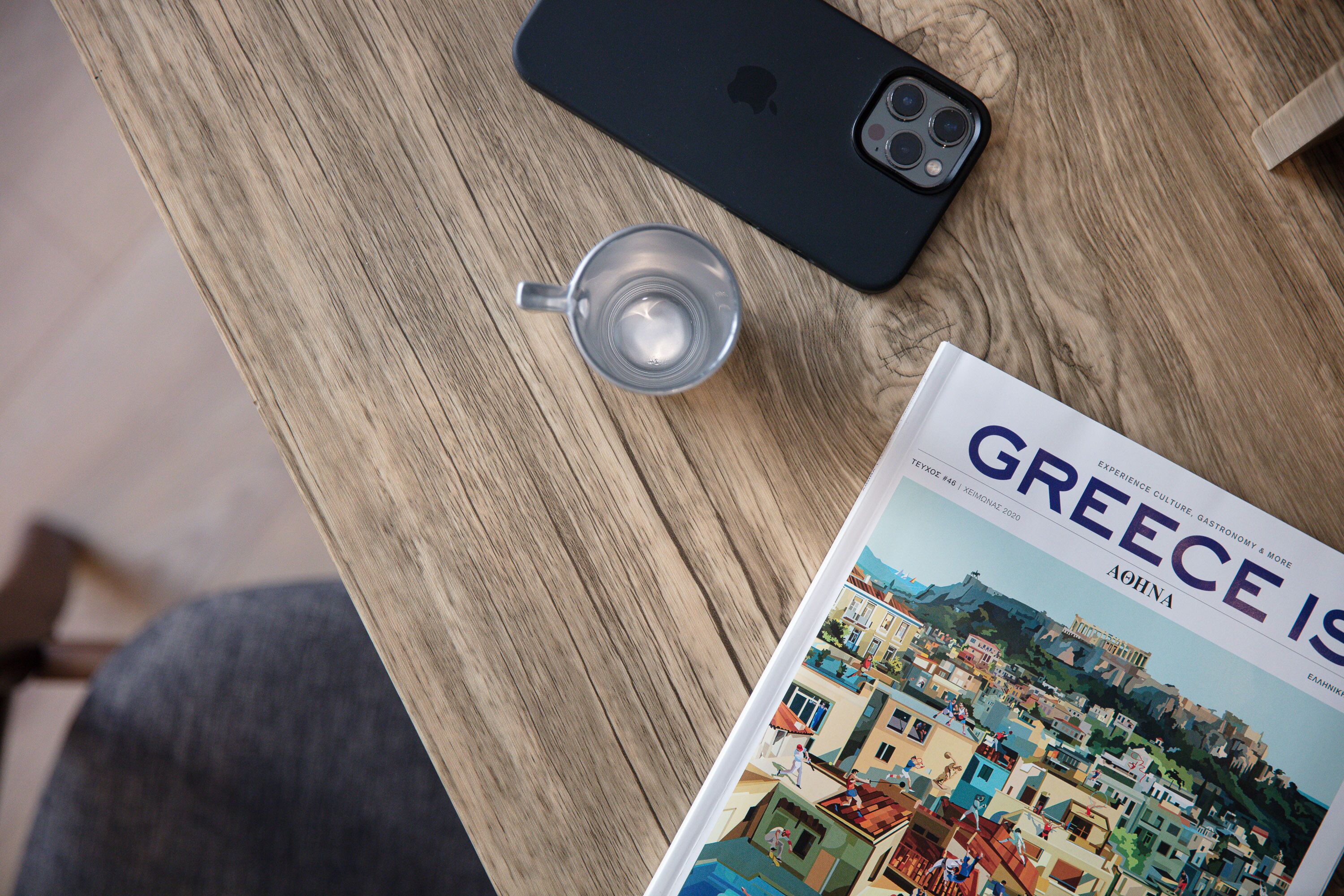 A magazine titled "Greece is..." on a wooden table with a smartphone and a glass of water.