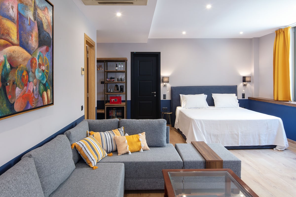 Une chambre d’hôtel avec un canapé gris, un lit avec du linge de lit blanc, une table basse en bois et une peinture abstraite colorée accrochée au mur.