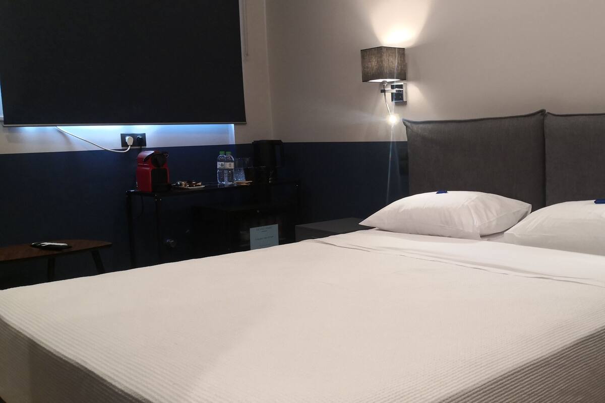 Une chambre d’hôtel avec un grand lit, deux oreillers, une table de chevet avec une lampe et une petite table sur laquelle se trouvent une machine à café et une bouteille d’eau.