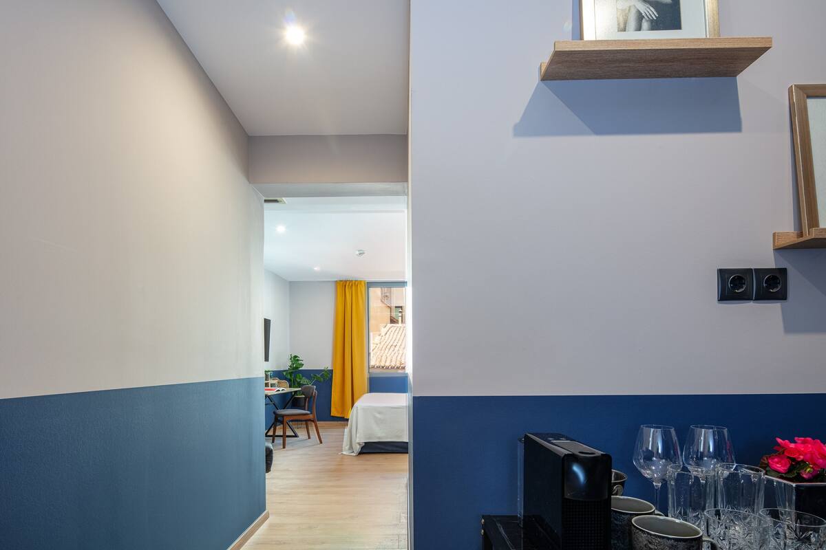 Une chambre d’hôtel moderne aux couleurs bleu et blanc, un coin repas avec des rideaux jaunes et un bar avec des verres et un téléviseur.