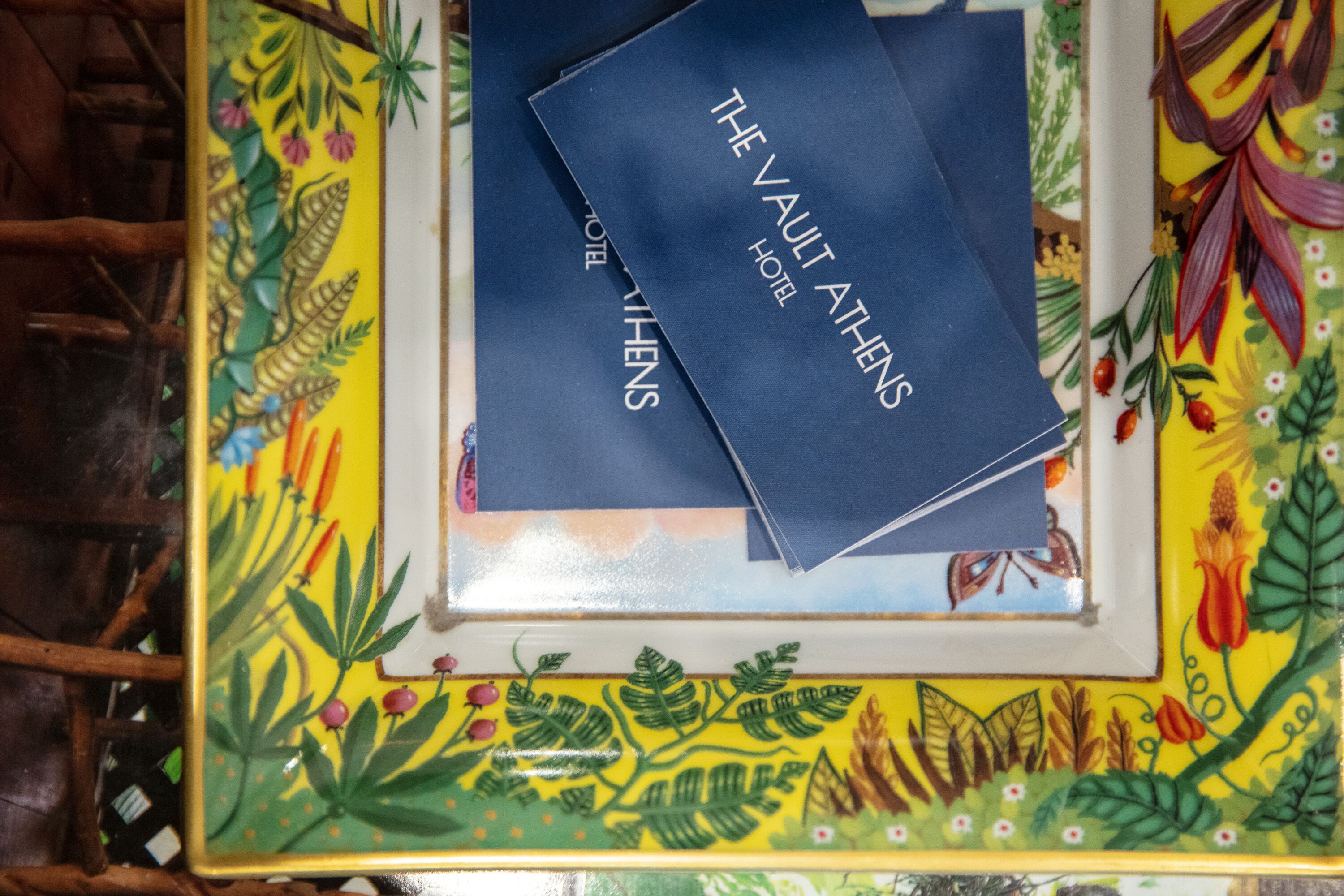 Un plateau décoratif orné d’un motif floral coloré et accompagné de deux livrets bleus intitulés « The Vault Athens Hotel ».