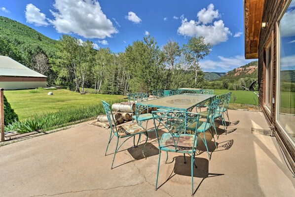 A patio with a table and chairs, a view of a green landscape, and a building with a glass door.