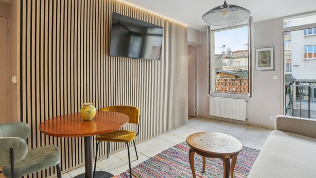A modern living room with a wooden slatted wall, a flat-screen TV, a round wooden table with a yellow vase, a yellow chair, a blue chair, a white sofa, and a colorful rug. - Hôtel Robespierre (Montreuil)