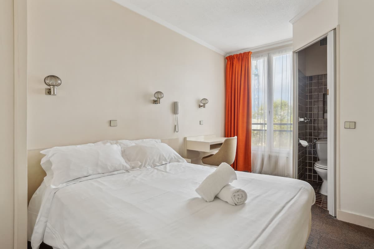 A hotel room with a bed, a desk, a telephone, and a view of the outside through a window with orange curtains.