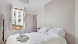 A neatly made bed with white linens and a rolled towel, a bedside table, and a window with a view of a building. - Hôtel Robespierre (Montreuil)