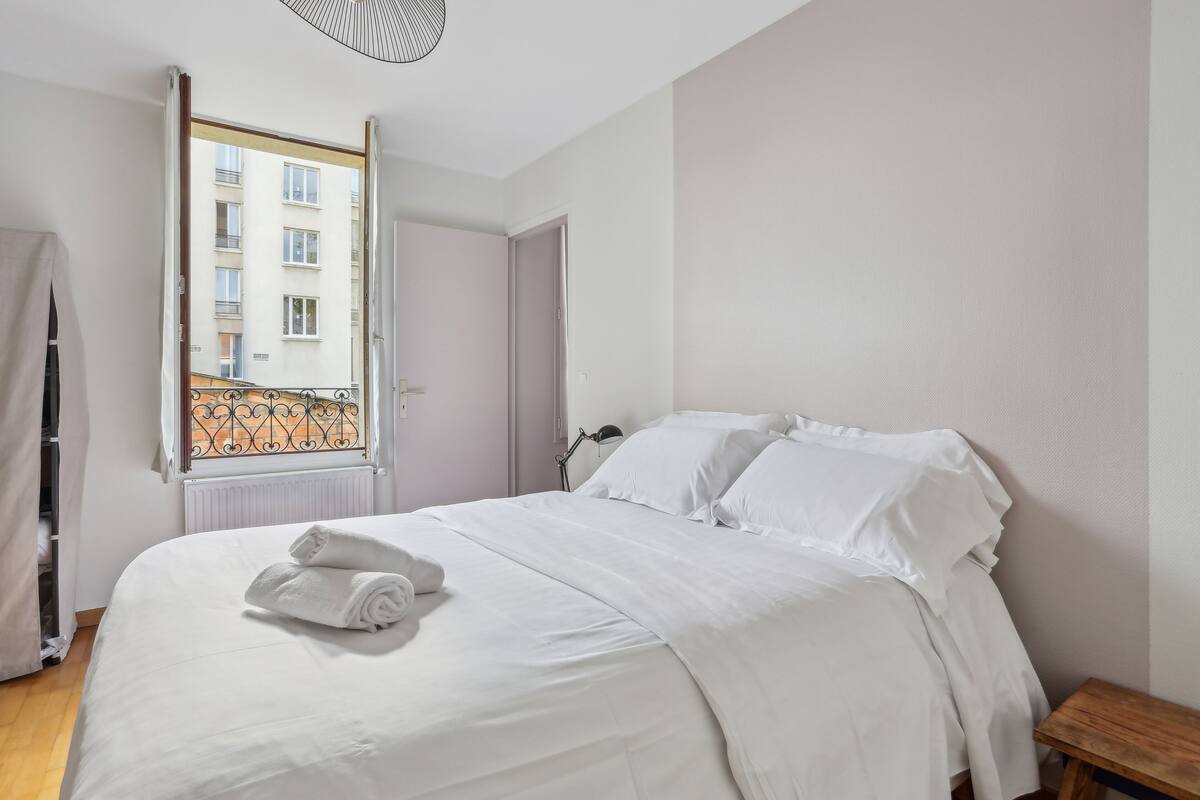 A neatly made bed with white linens and a rolled towel, a bedside table, and a window with a view of a building.