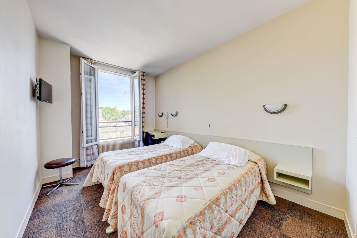 A hotel room with two beds, a small stool, a window with a view, and a wall-mounted shelf.