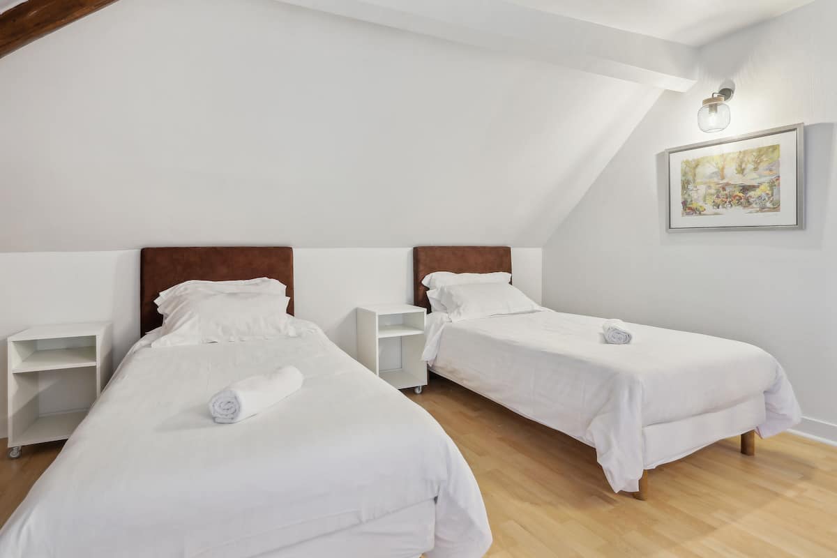 A room with two beds, white bedding, wooden headboards, a white nightstand, a small shelf, and a painting on the wall.