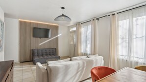 A modern living room with a sectional sofa, a grey armchair, a wooden dining table, and a TV mounted on a wall with vertical blinds. - Hôtel Robespierre (Montreuil)