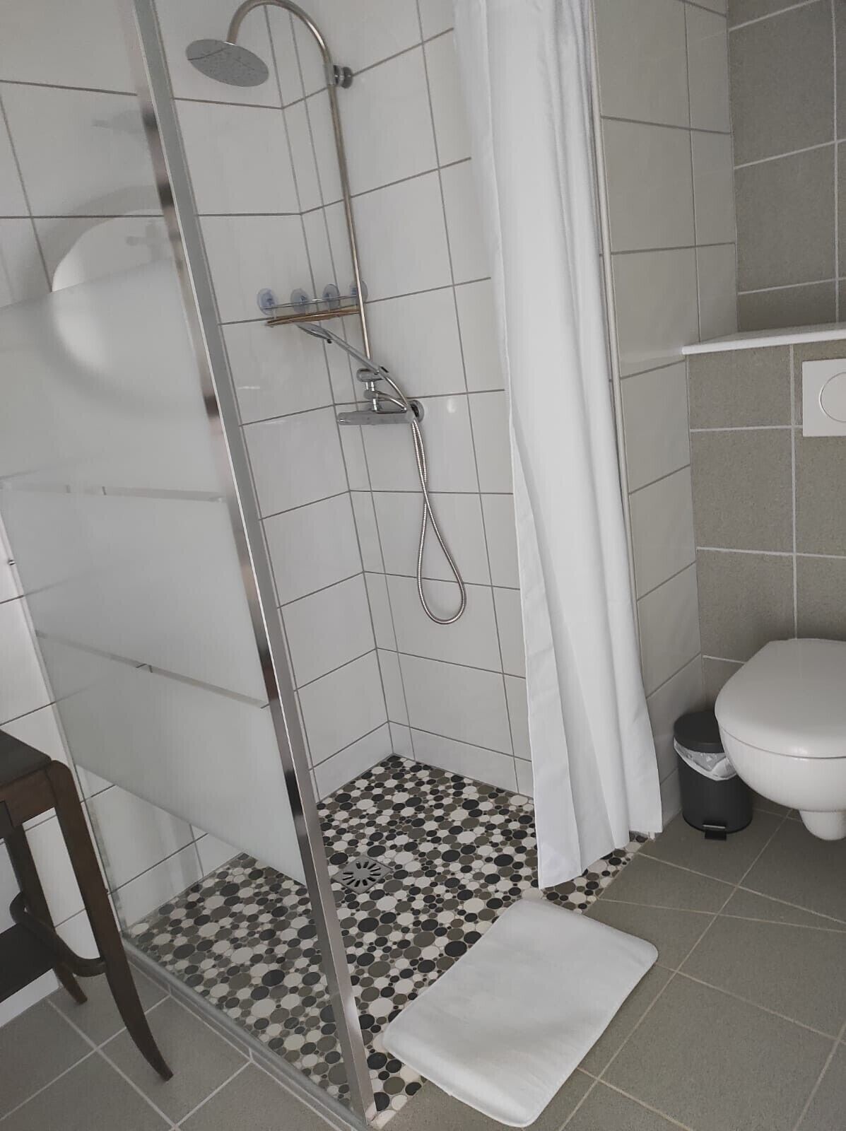 Suihkukaappi, jossa on lasiovi, valkoinen verho ja geometrisesti kuvioitu laattalattia.