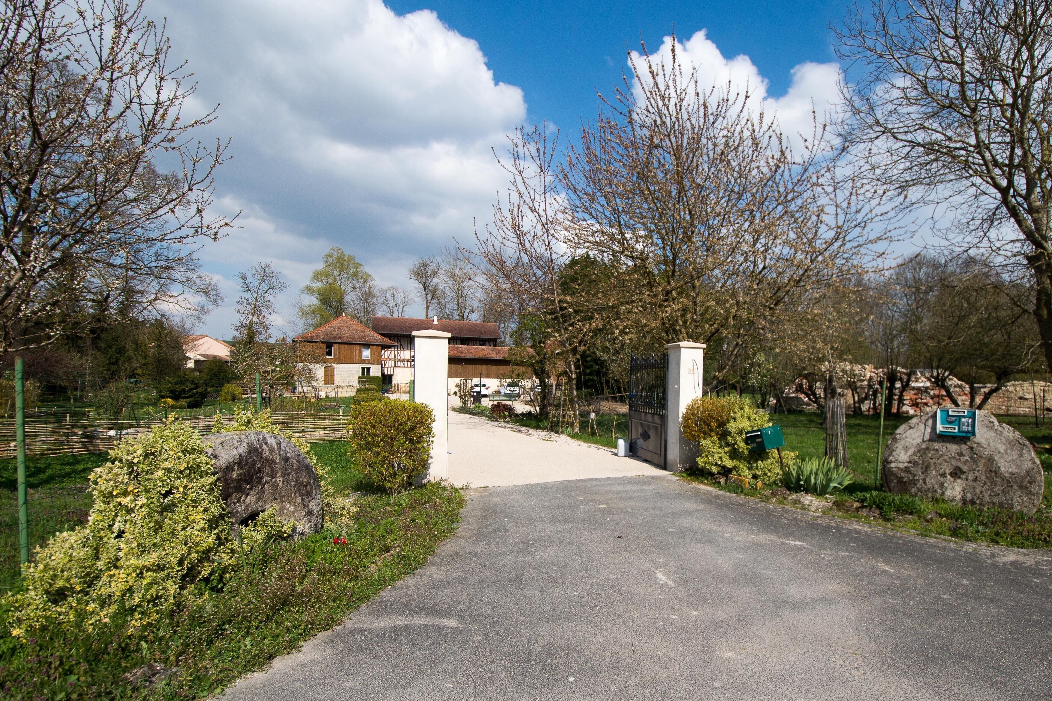 Une entrée fermée par un portail donne sur une allée pavée qui mène à un bâtiment doté d’un toit rouge, entouré de verdure et d’arbres.