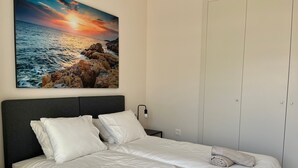 A neatly made bed with white linens and rolled towels, a nightstand with a lamp, and a painting of a sunset over the ocean on the wall. - Apartment Lotus 486 with Shared Pool, Terrace & Wi-Fi (LA CALA DE MIJAS)
