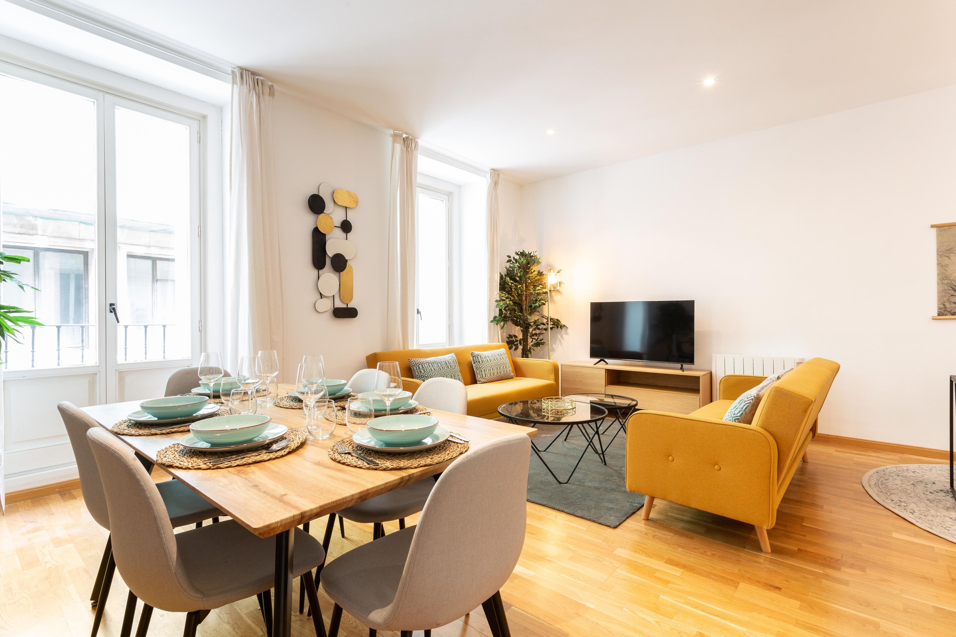 Una sala moderna con una mesa de comedor de madera para cuatro personas, un sofá, un televisor y una planta.