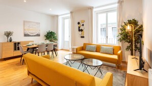 Ein modernes Wohnzimmer mit einem gelben Sofa, einer Essecke mit Tisch und Stühlen sowie einem großen Fenster mit Vorhängen.