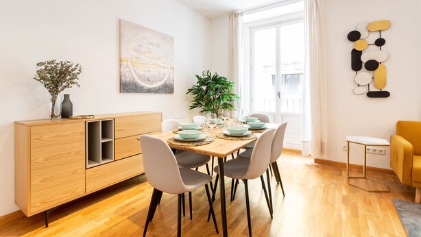 Ein Essbereich mit einem Holztisch, gedeckt für vier Personen, umgeben von grauen Stühlen. Eine Holzkommode mit einer Vase und einer Pflanze steht links.