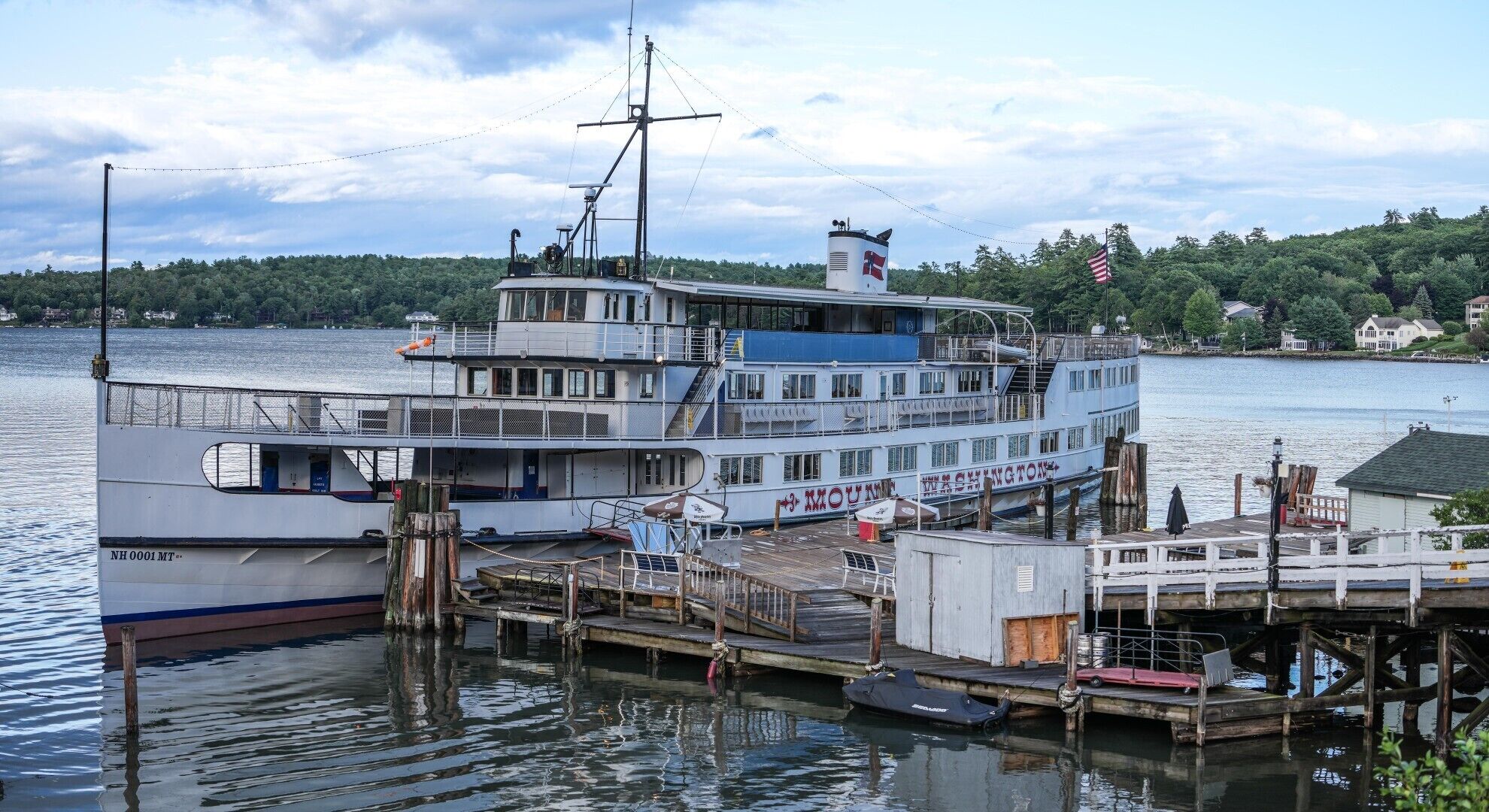 A large multi-deck boat docked at a pier with a cabin structure, a lifeboat, and a flag.