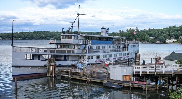 A large multi-deck boat docked at a pier with a cabin structure, a lifeboat, and a flag.