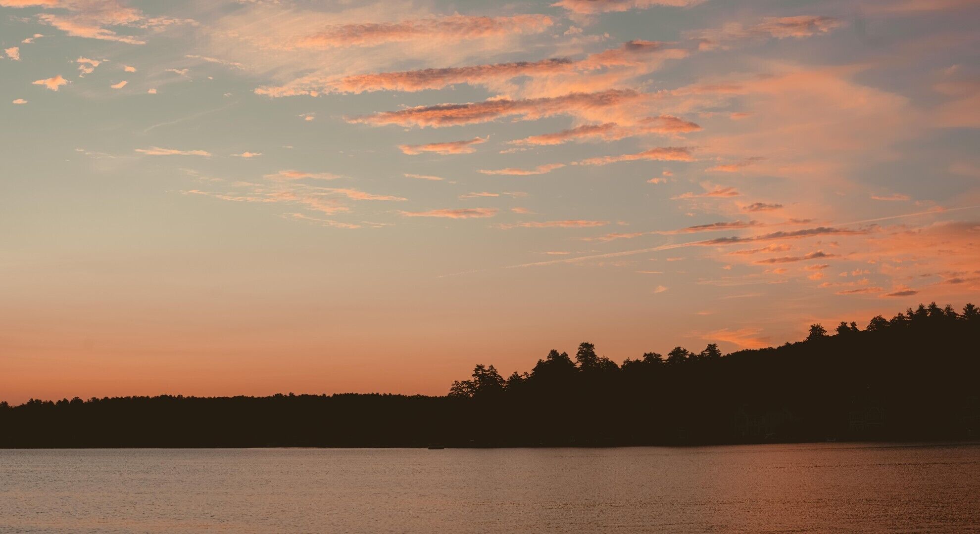 A sunset over a calm body of water with a silhouette of a forested hill in the distance.