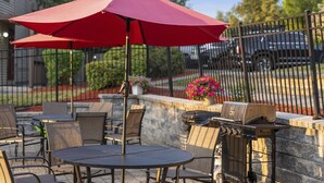 Un espacio de patio al aire libre con una mesa redonda, sillas, una parrilla y un paraguas rojo.