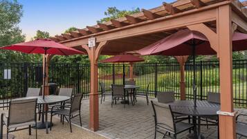 Un espace extérieur aménagé en terrasse, avec des tables et des chaises, recouvert de pergolas en bois et ombragé par des parasols rouges.