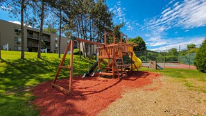Children can play on the playground's slide and swing set amidst trees and a secure fence.