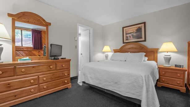 Ein Hotelzimmer mit einem Bett, einem Holzschrank, einem Fernseher, zwei Nachttischlampen, einem Bild an der Wand und einem Fenster mit Vorhängen.