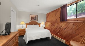 Una habitación de hotel con cama, dos lámparas de noche, un televisor sobre un aparador de madera y una ventana con cortinas.