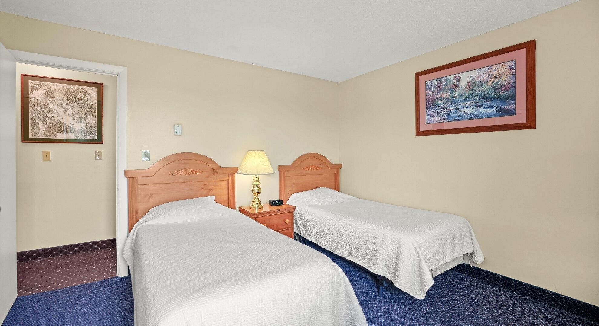 Camera d'albergo con due letti singoli, un comodino con una lampada, un quadro appeso e una porta.