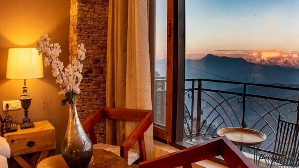 A room with a view of mountains, a wooden table with a vase of flowers, a lamp, and a chair. - The Himalayan Voyage (Ranikhet)