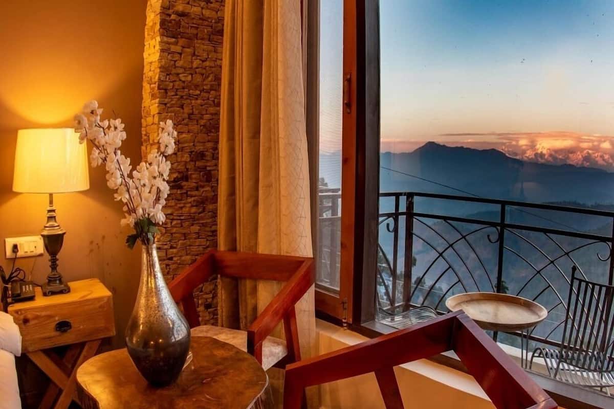 A room with a view of mountains, a wooden table with a vase of flowers, a lamp, and a chair.