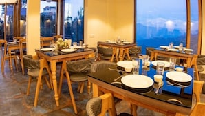 A dining area with wooden tables and chairs, large windows with a view, and pendant lighting. - The Himalayan Voyage (Ranikhet)