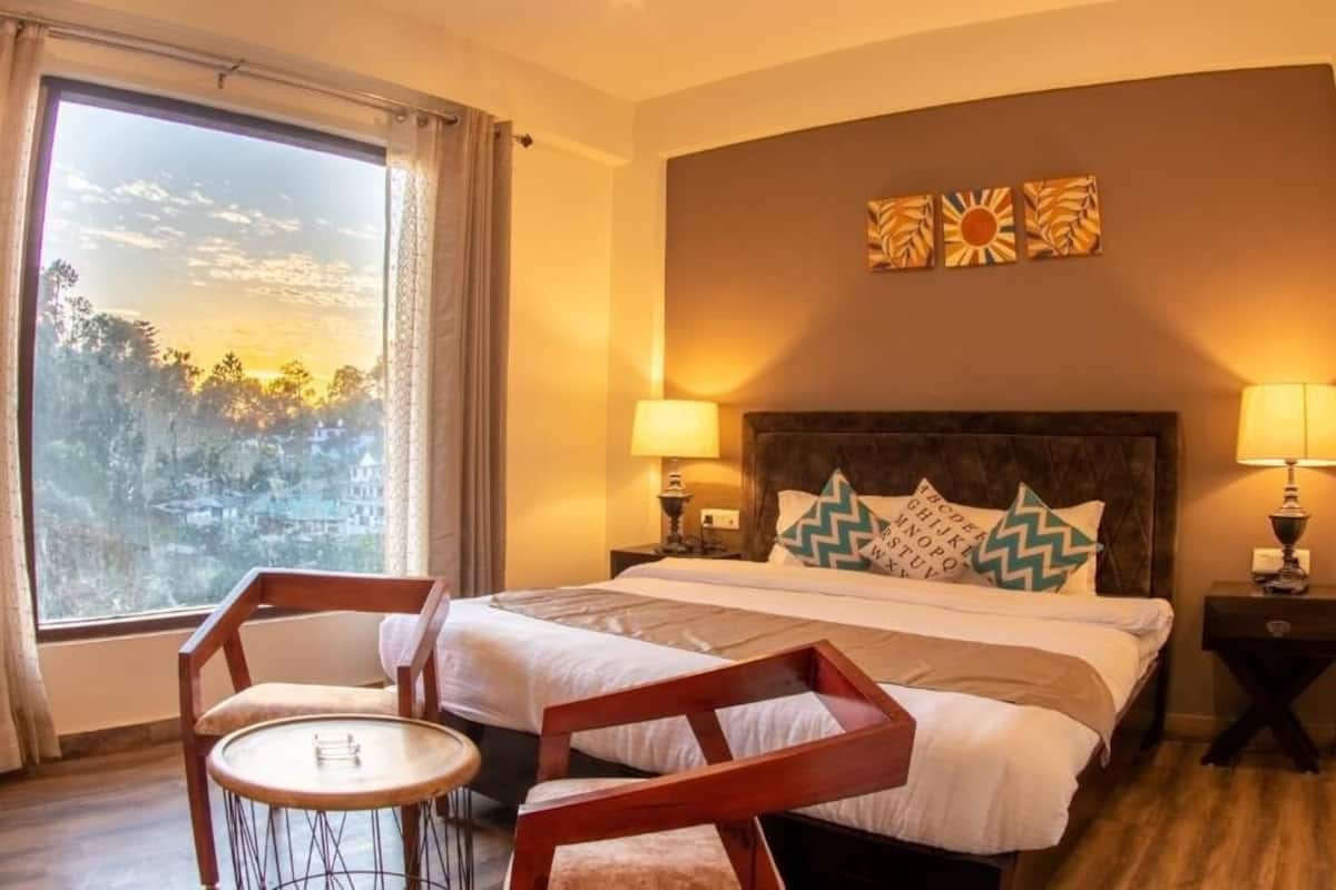 A hotel room with a bed, two bedside tables with lamps, a chair, a small round table, and a large window with curtains.