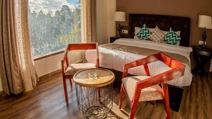 A bedroom with a bed, two chairs, a small round table, and a view of a forest through the window. - The Himalayan Voyage (Ranikhet)