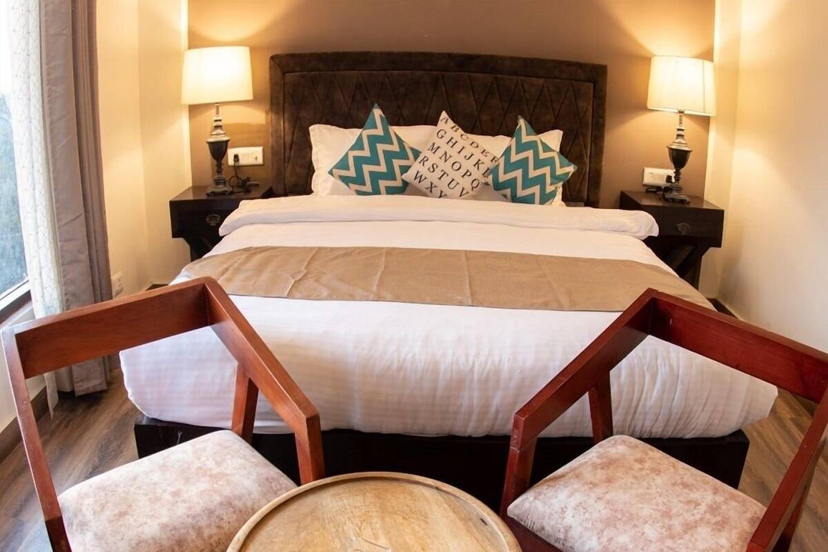 A hotel room with a large bed, two nightstands with lamps, a wooden side table, and two upholstered chairs.